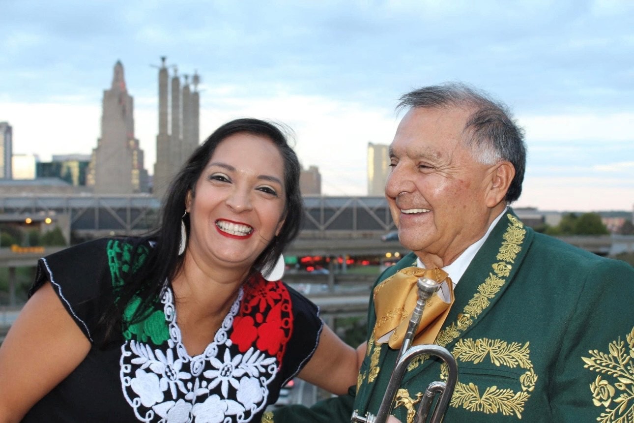 A young woman and older man smile into the camera with a city skyline obscured in the background.