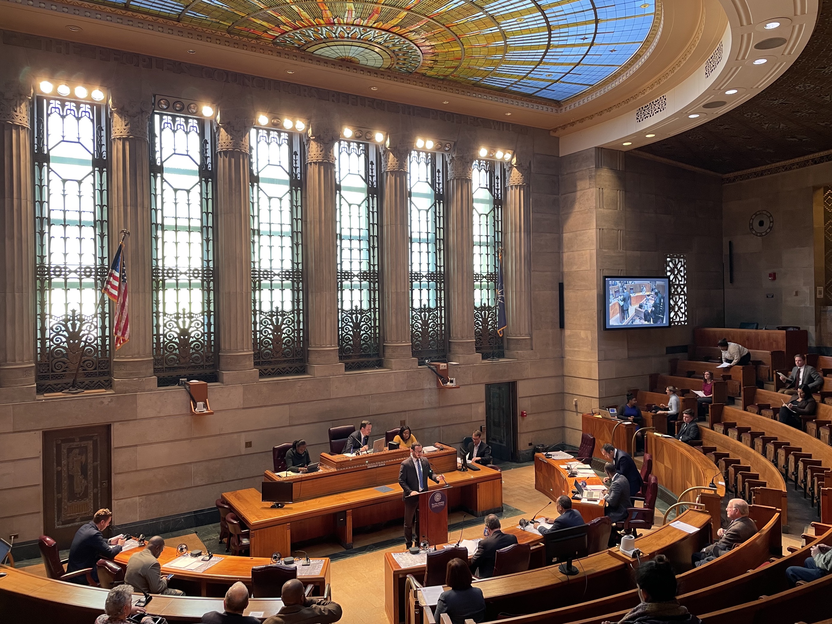 The inside of the Buffalo Common Council chambers is shown in a wide angle. The seating area of the auditorium is brown wood, with people seated facing the front where the chair person is sat facing outward. Councilmember Mitch Nowakowski stands at a brown lectern at the front of the chambers facing outward. He is wearing a dark suit with a blue tie.