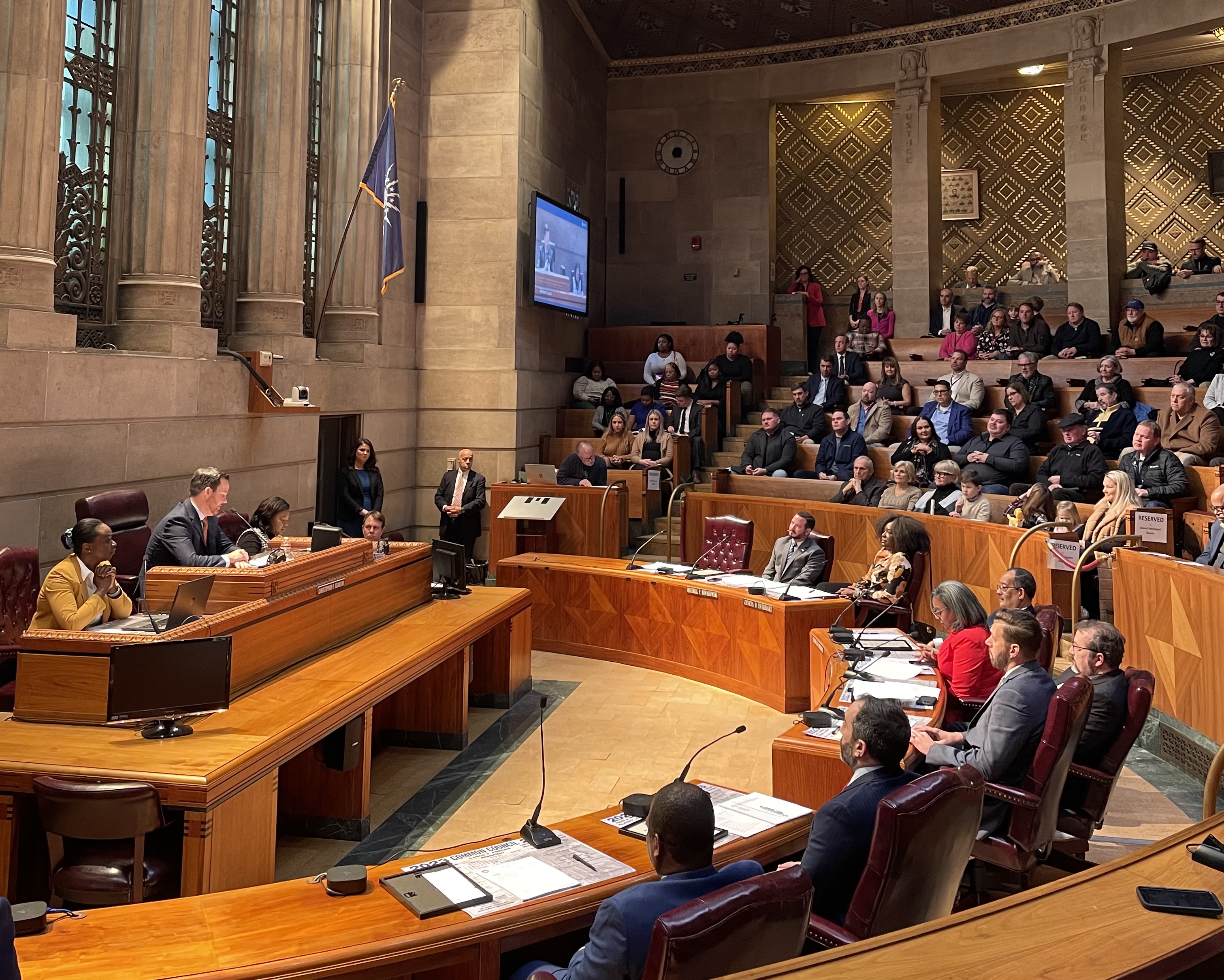 The common council chambers is pictured in a wide shot. People are gathered in the chamber and councilmembers are seated in red leather chairs. They are facing Christopher P. Scanlon, seated in the President's chair.