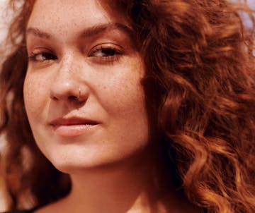 Foto de uma mulher jovem, branca e de cabelos cacheados, com um sorriso no rosto, vestindo agasalho verde musgo e camiseta verde água, mexendo em um smartphone. Ela está segurando o aparelho com as duas mãos. Ao fundo, tem-se a fachada de um prédio e diversas plantas compostas por folhas verdes.