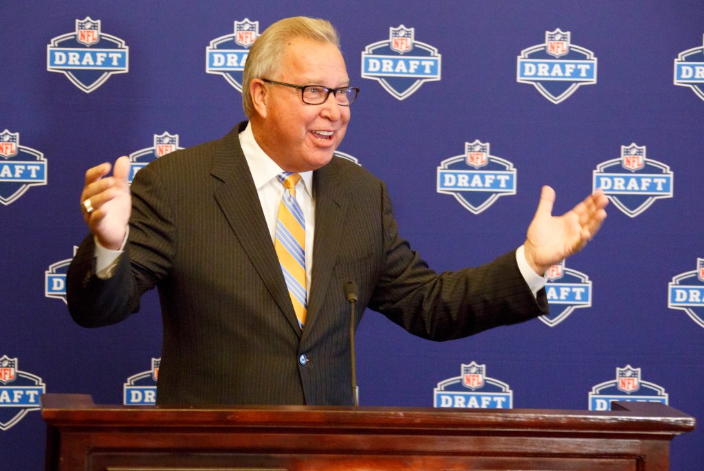 Ron Jaworski speaking at a press conference in front of a blue wall with logos and journalists surrounding him.