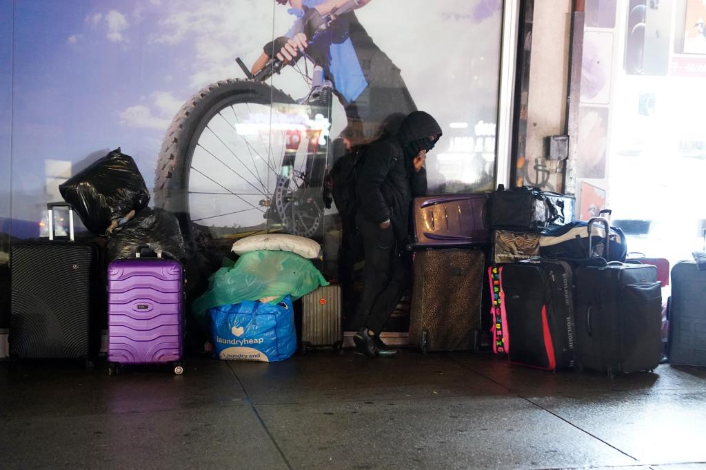 Man stands along suitcases piled against a wall on the sidewalk. 