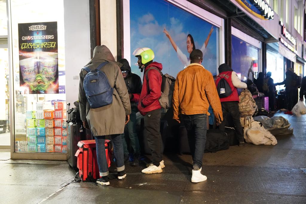 Multiple people stand with their suitcases on sidewalk in the rain. 