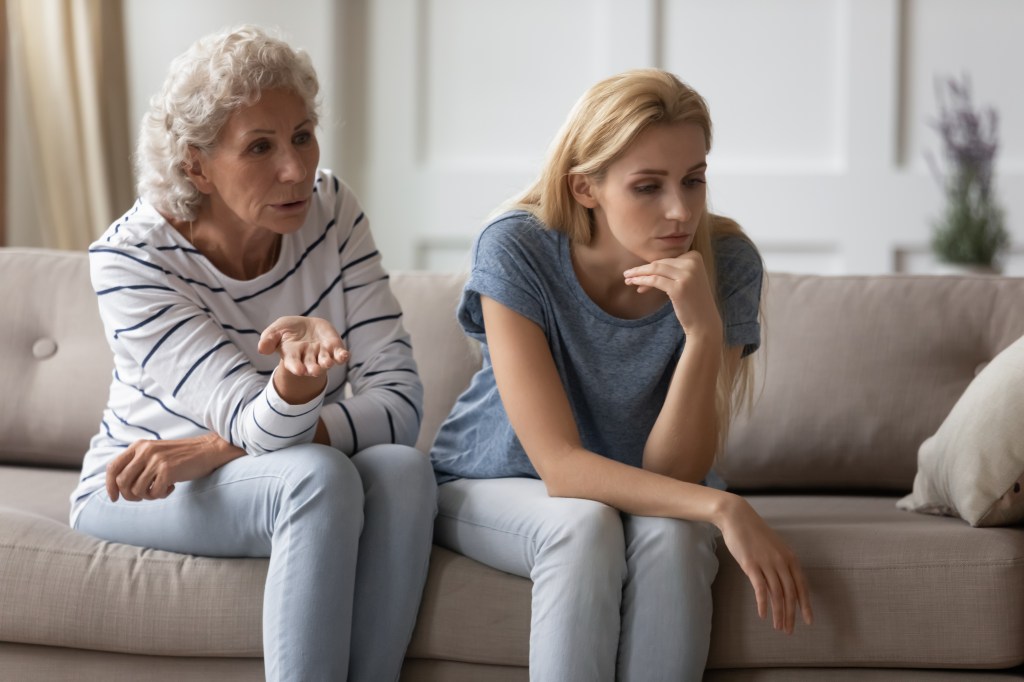 Unhappy young woman sitting on sofa, trying to avoid conflict with scolding older woman