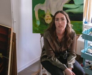 Portrait of a yound woman in her painting studio with a canavs behind.