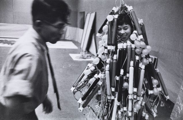 A black-and-white photograph of Tanaka Atsuko’s "Electric Dress," showcasing the artist wearing an intricate, bulb-covered costume of neon tubes and light bulbs, emphasizing experimental and kinetic art.