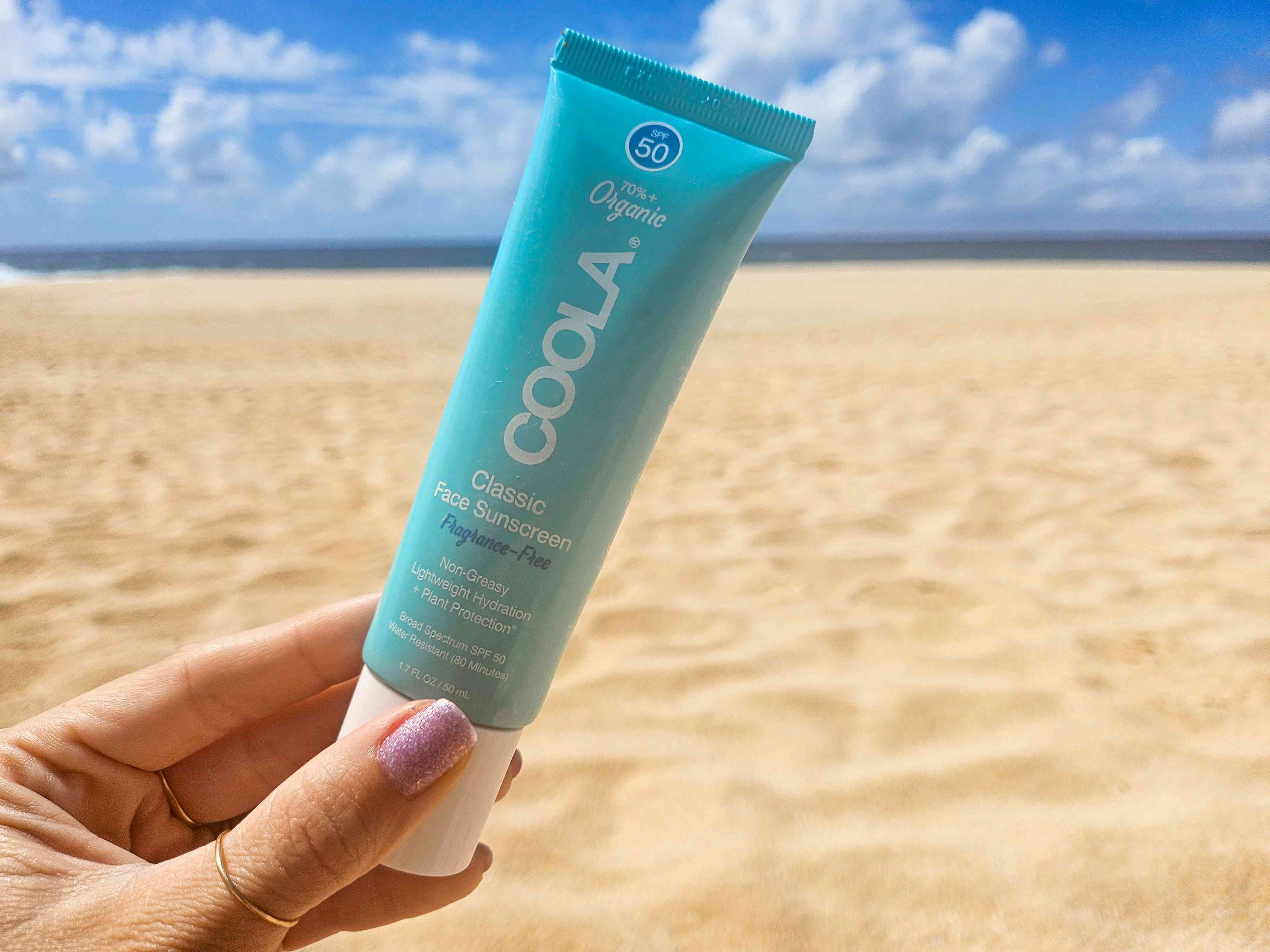 Hand holding the Coola Organic Face Sunscreen with sand and blue sky in background