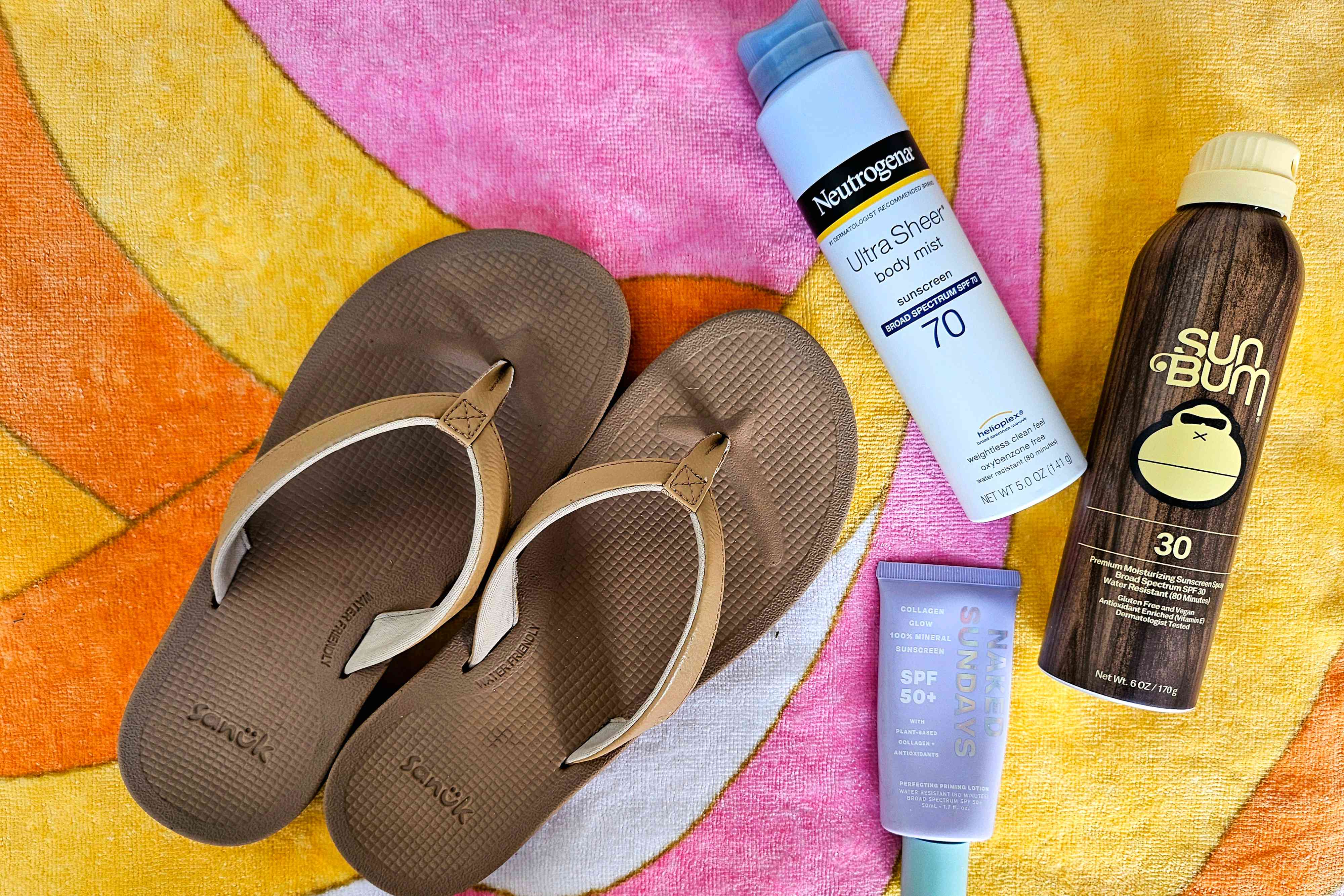 Sanuk Cosmic Shores Flip Flops next to three sunscreen products on a colorful beach towel