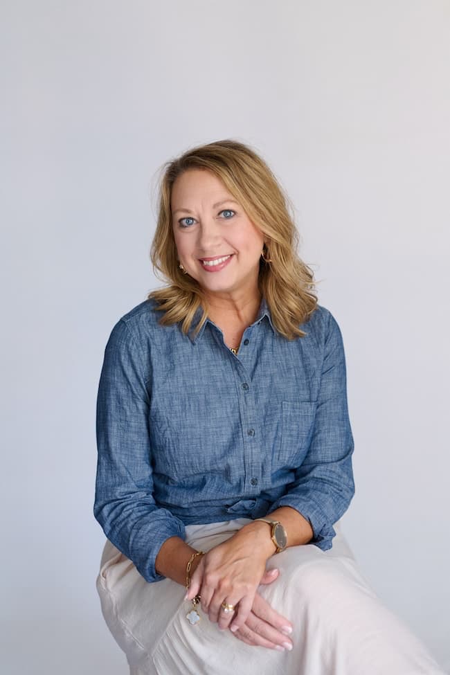 A woman with shoulder-length blonde hair smiles while sitting against a plain white background. She is wearing a blue button-up shirt and light-colored pants. She has her hands folded on her lap and is looking directly at the camera.