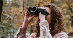 A person with curly hair wearing a plaid shirt is looking through binoculars. They are standing outdoors with a blurred background of autumn foliage.