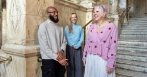 Three people stand in a marble staircase area, smiling and engaged in conversation. The person on the left wears glasses and a sweater, the person in the middle wears a blue sweater, and the person on the right wears a pink polka dot top.