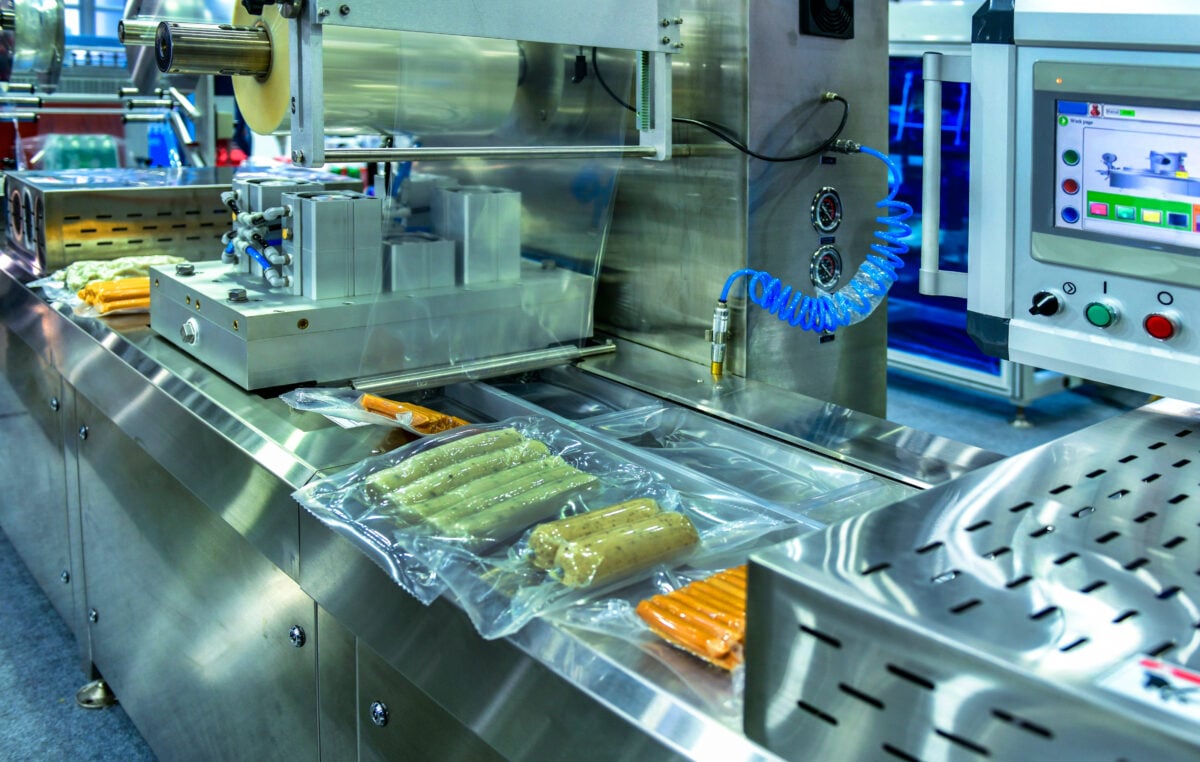 Photo shows a silver food-processing machine with some cheese, sausages, and other items on top