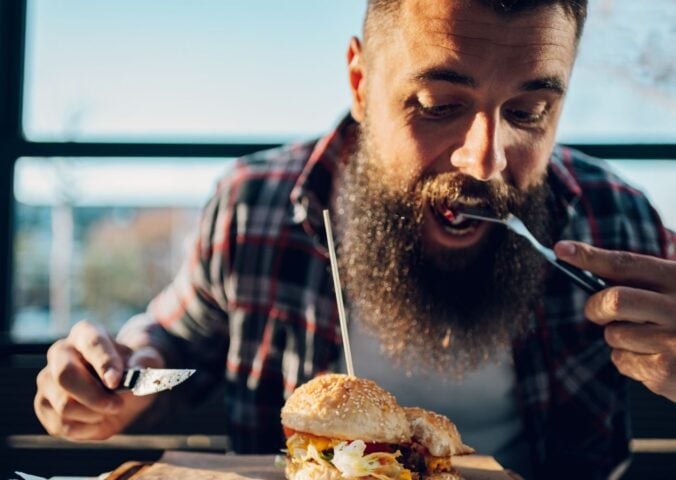 Photo shows a bearded man eating a burger with a knife and fork