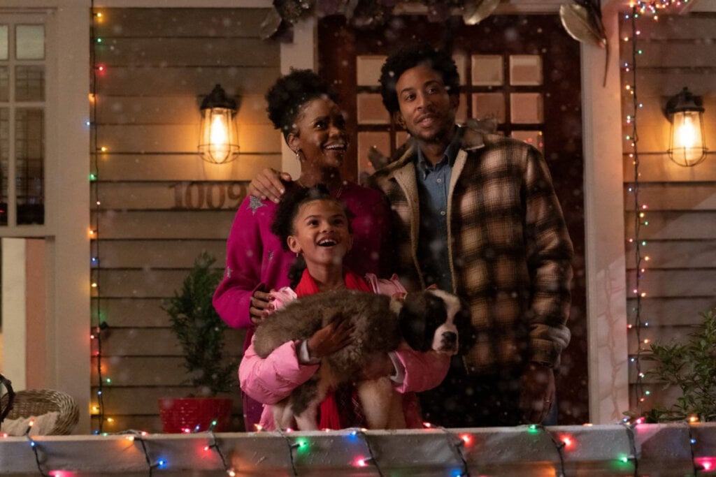 Casal na porta de casa com a filha e um cachorro observando a neve cair 