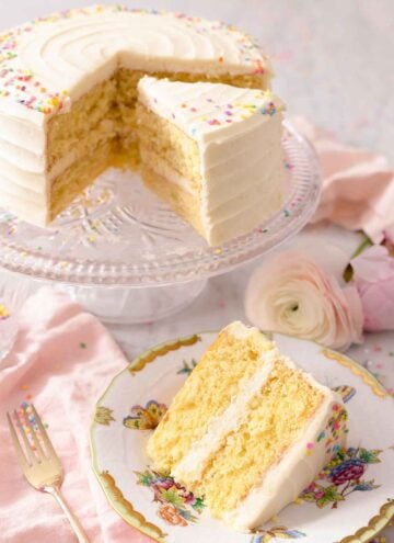 A slice of vanilla cake on a plate in front of the cake on a cake stand.