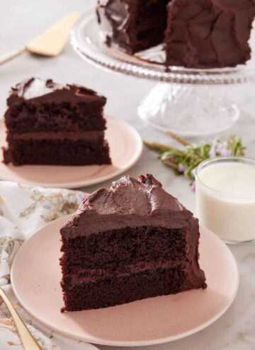 Two plates with sliced chocolate cake along with a glass of milk and cake stand with more cake.