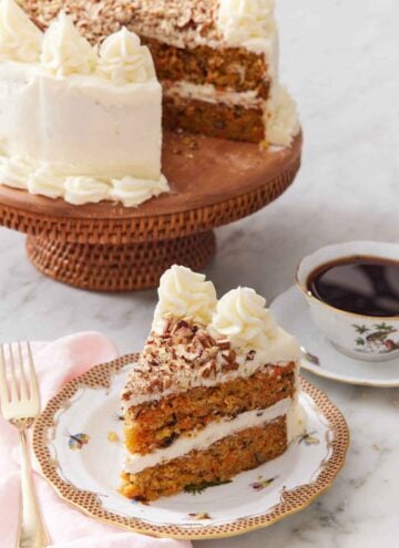 A slice of carrot cake with the rest of the cake on a cake stand in the back along with a cup of coffee.