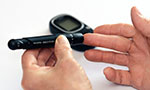 Photo: a hand holding a glucose meter and pointing to a finger of the other hand. In the background, a glucose monitor.