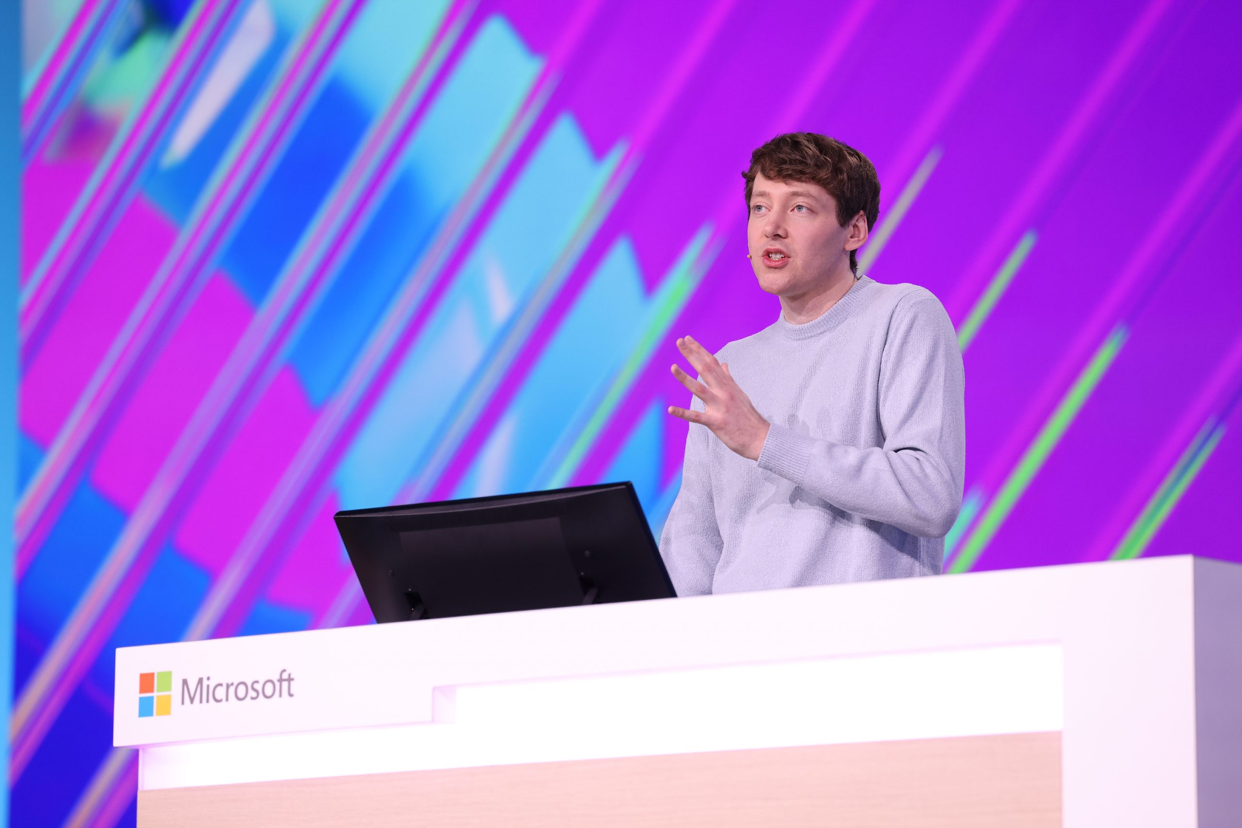 A man standing on stage behind a table with a laptop on it and a multi-colored screen behind him