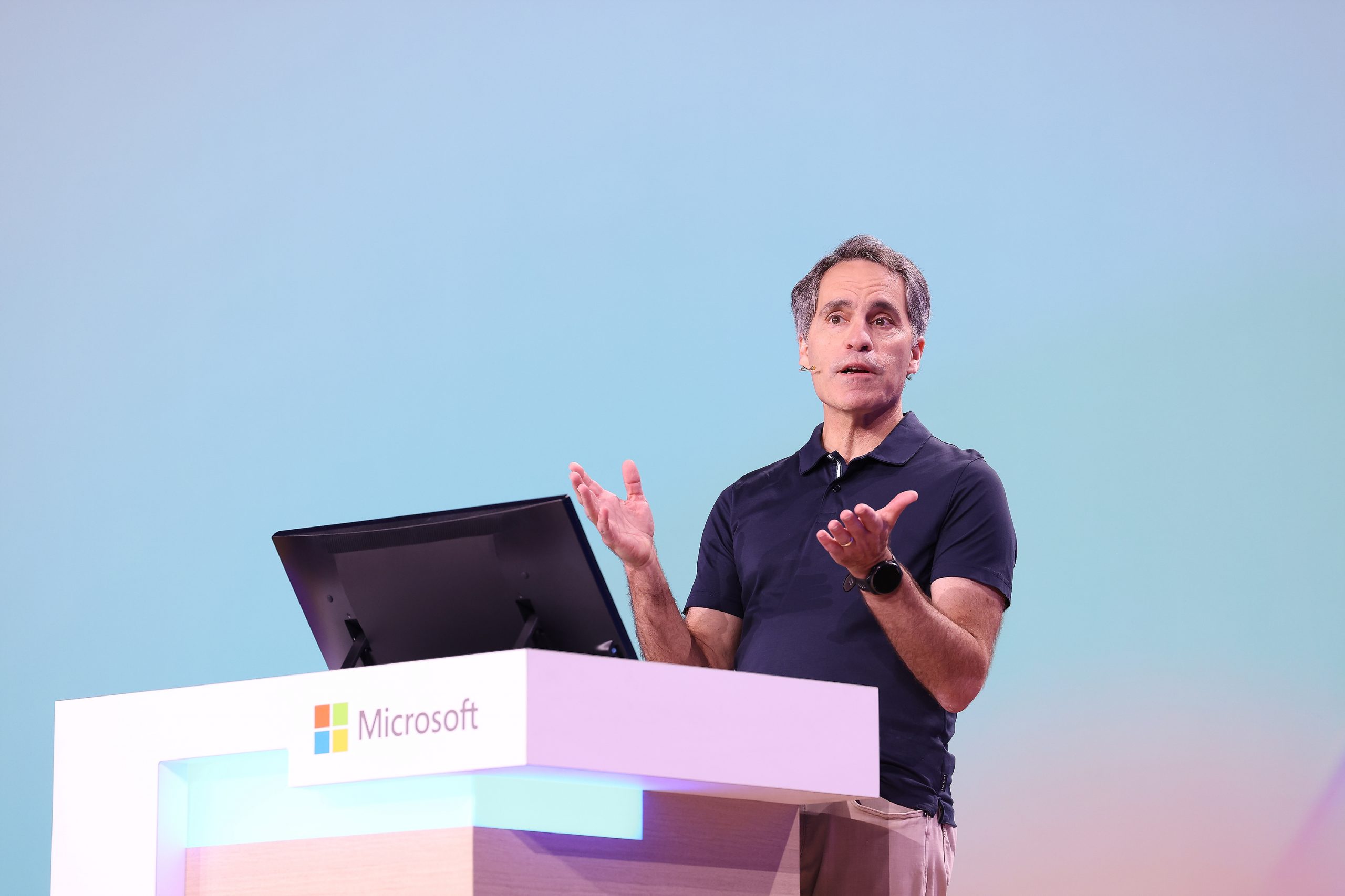 A man standing on stage with a table and laptop in front of him and a blue and rose-gold screen behind him