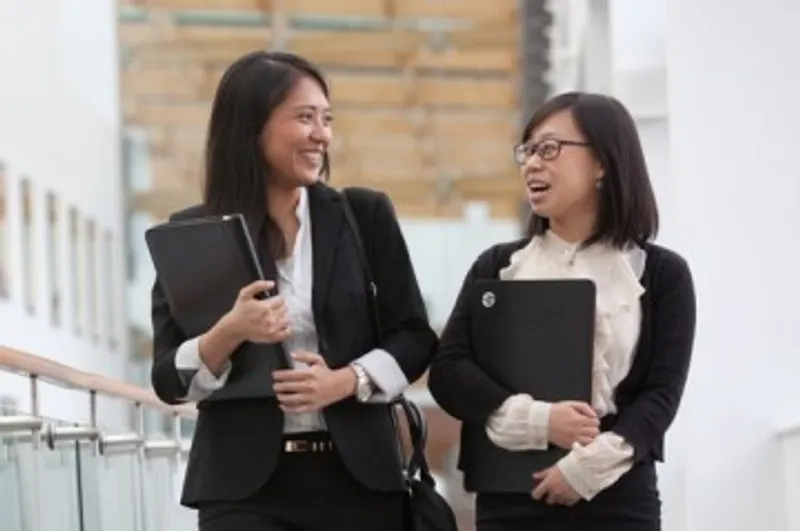 Two businesswomen carrying laptops and talking while walking along a corridor