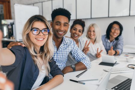 African and european girls posing with peace sign while blonde lady making selfie in conference hall. Young african web-developer having fun with colleagues and smiling for photo.