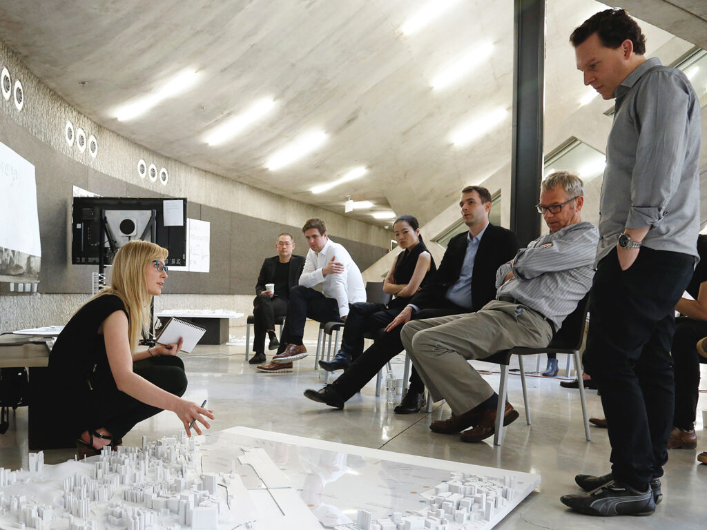 A woman crouches next to a model and speaks to a group of onlookers.