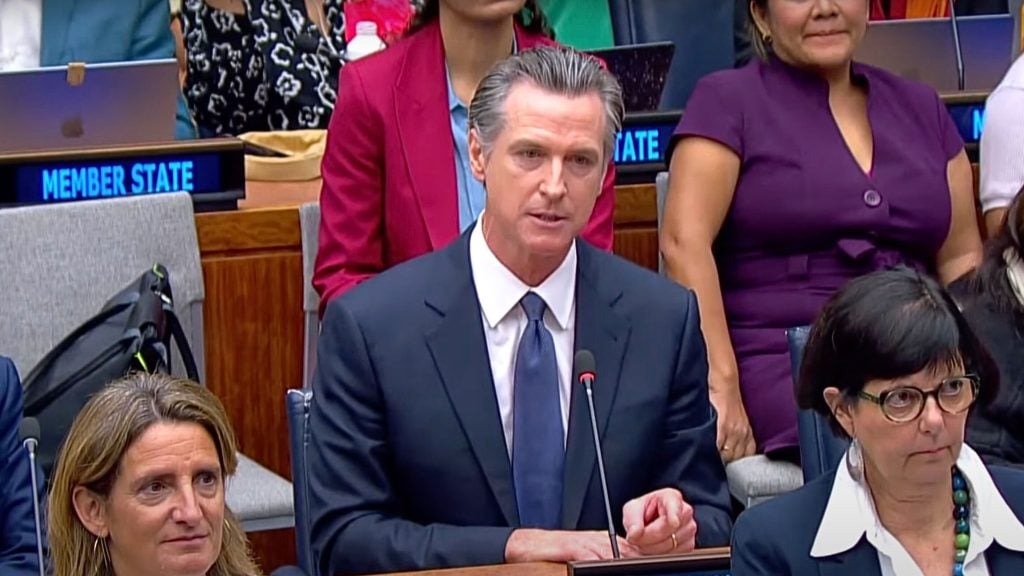 A person in a suit and tie speaks into a microphone at a formal meeting, while others sit and listen in the background.