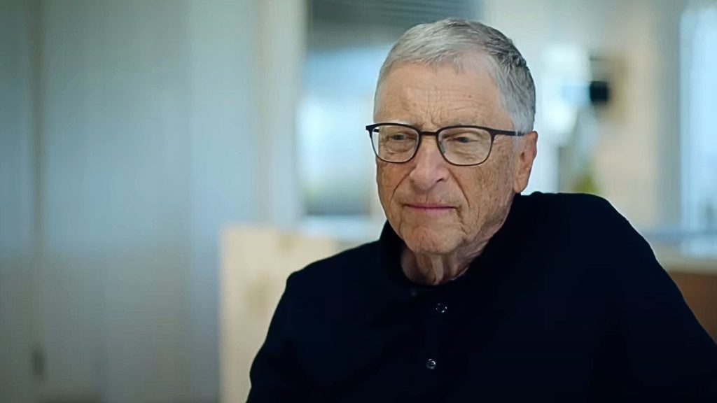 Bill Gates with short gray hair and glasses wearing a black shirt, sitting in a well-lit indoor setting.