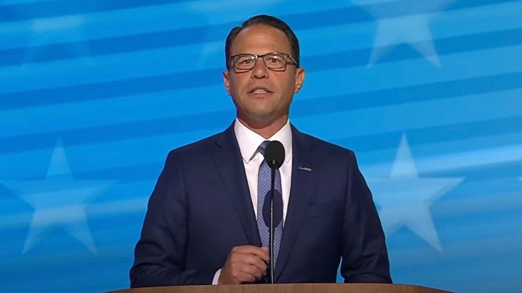 Gov. Josh Shapiro and tie is speaking at a podium. The background is blue with white stars.