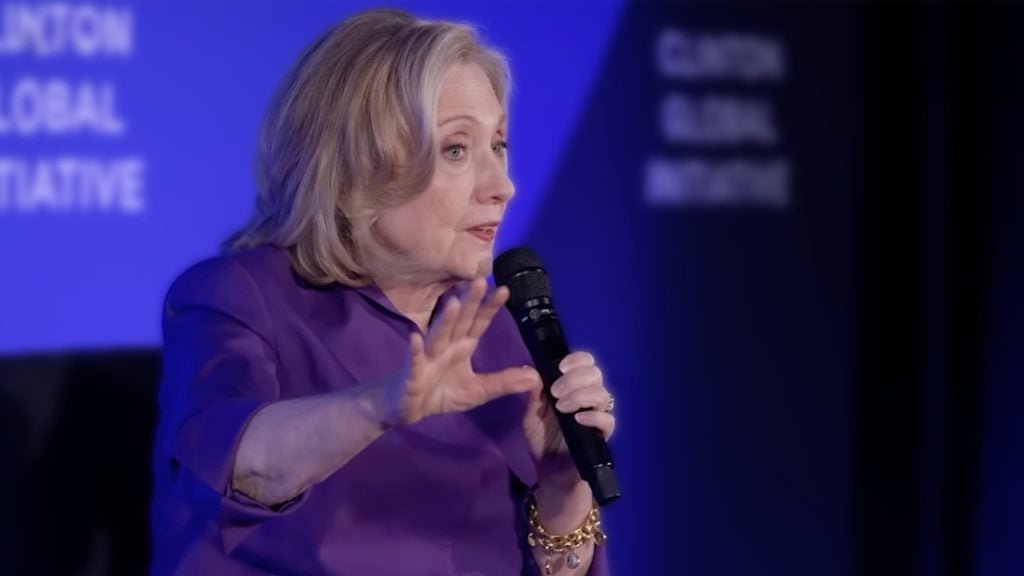 Hillary Clinton in a purple outfit speaks into a microphone against a blue background that reads "Clinton Global Initiative."