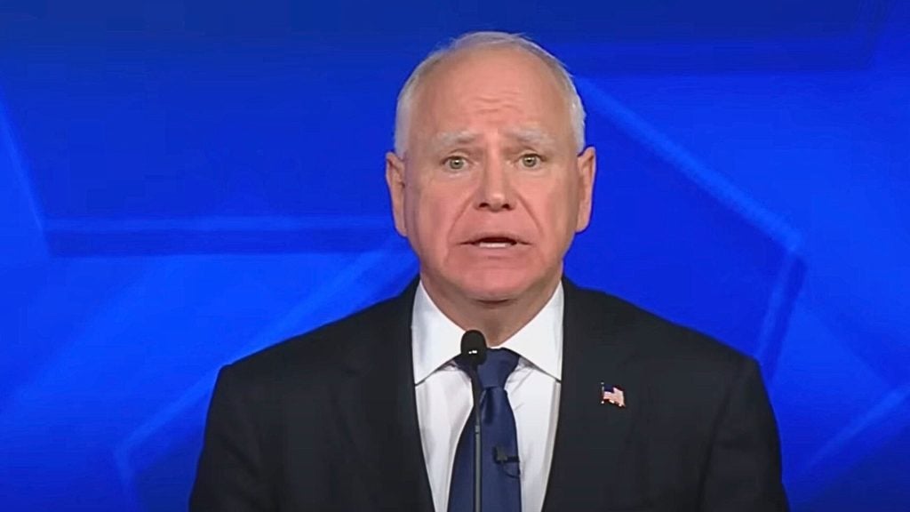 Tim Walz in a suit and tie speaking into a microphone with a blue background.