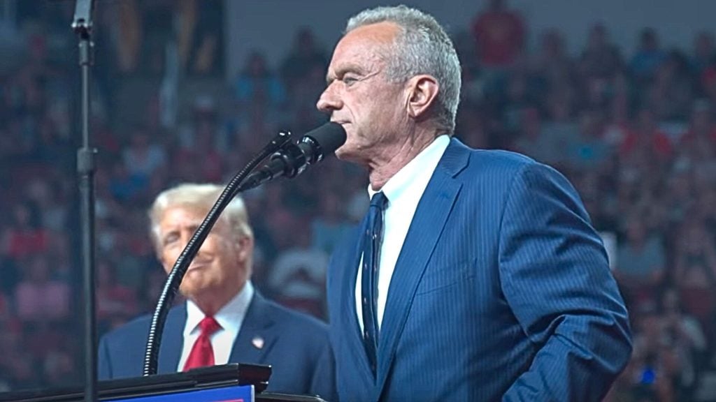 RFJ Jr. in a blue suit speaking at a podium with a microphone, with a blurred crowd in the background.