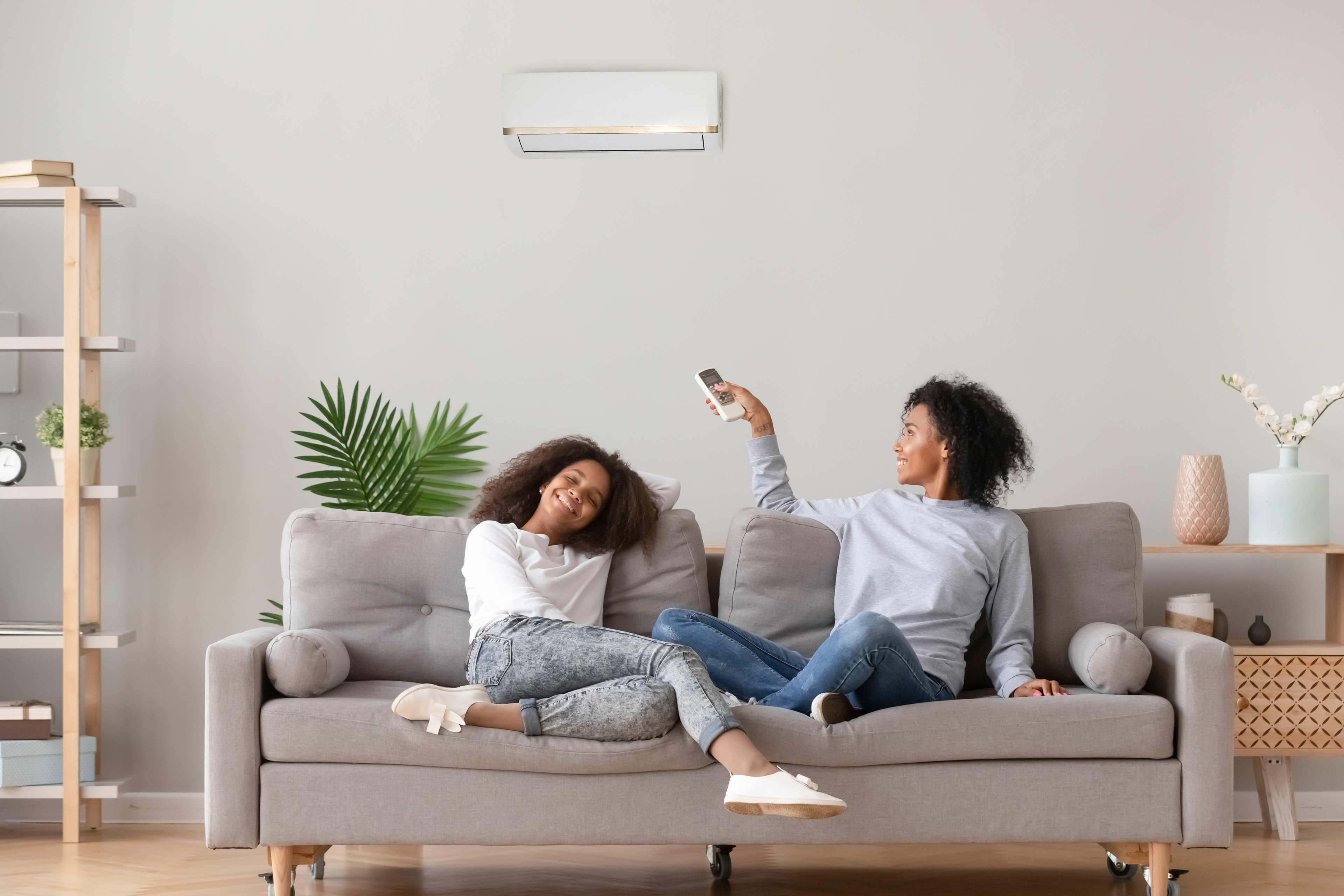 Woman and child sitting and smiling on a gray couch as woman points a remote at an air conditioning unit on the wall behind them