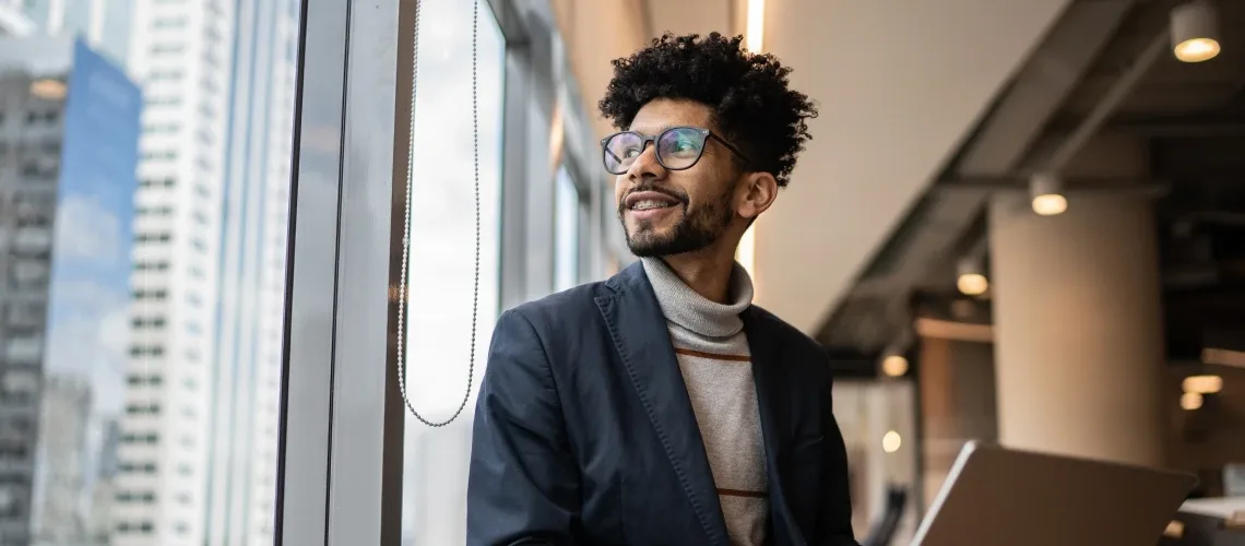 Pessoa de óculos e cabelo cacheado vestindo blusa de gola alta e blazer, sentada perto de uma janela com um notebook e olhando para fora de um escritório moderno.