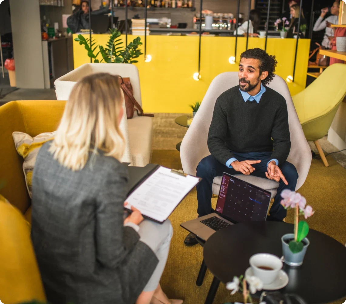 Dois indivíduos conversando no lounge de um escritório moderno. O homem está sentado em uma cadeira, gesticulando, enquanto a mulher segura uma prancheta. Um notebook e uma xícara de café estão na mesa entre eles.