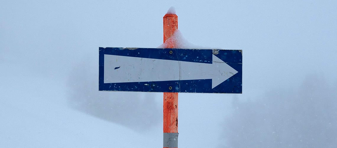 A snow-covered direction sign with a white arrow pointing to the right stands on a red and white pole against a foggy background.