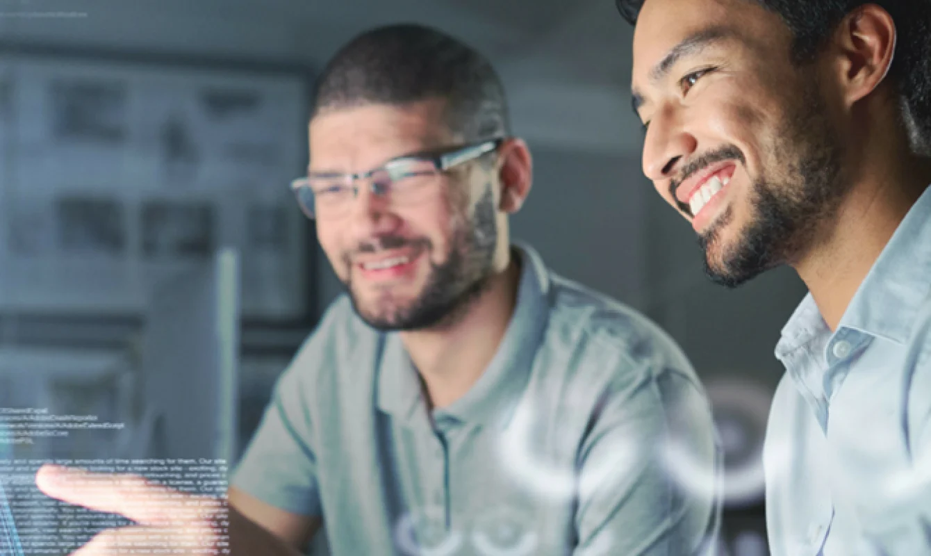 Two men are smiling and looking at a computer screen. One is pointing, and there is code visible on the screen. They appear to be in a collaborative work environment.