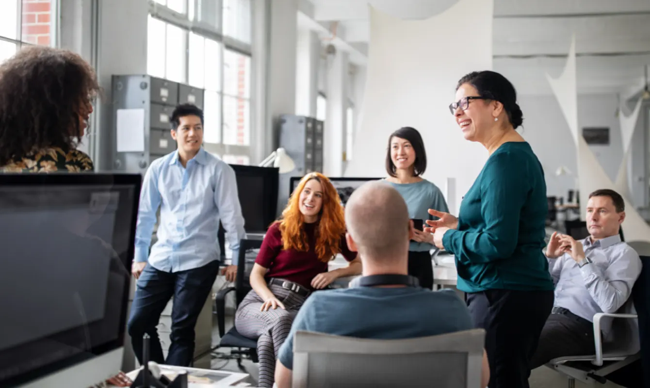 A group of six people sit and stand around a meeting space in an office, engaging in a lively conversation and smiling.