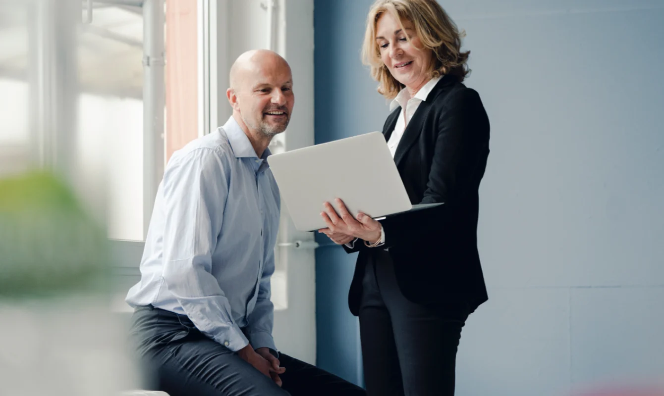 A man sits while a woman stands, both professionally dressed, looking at a laptop she is holding.