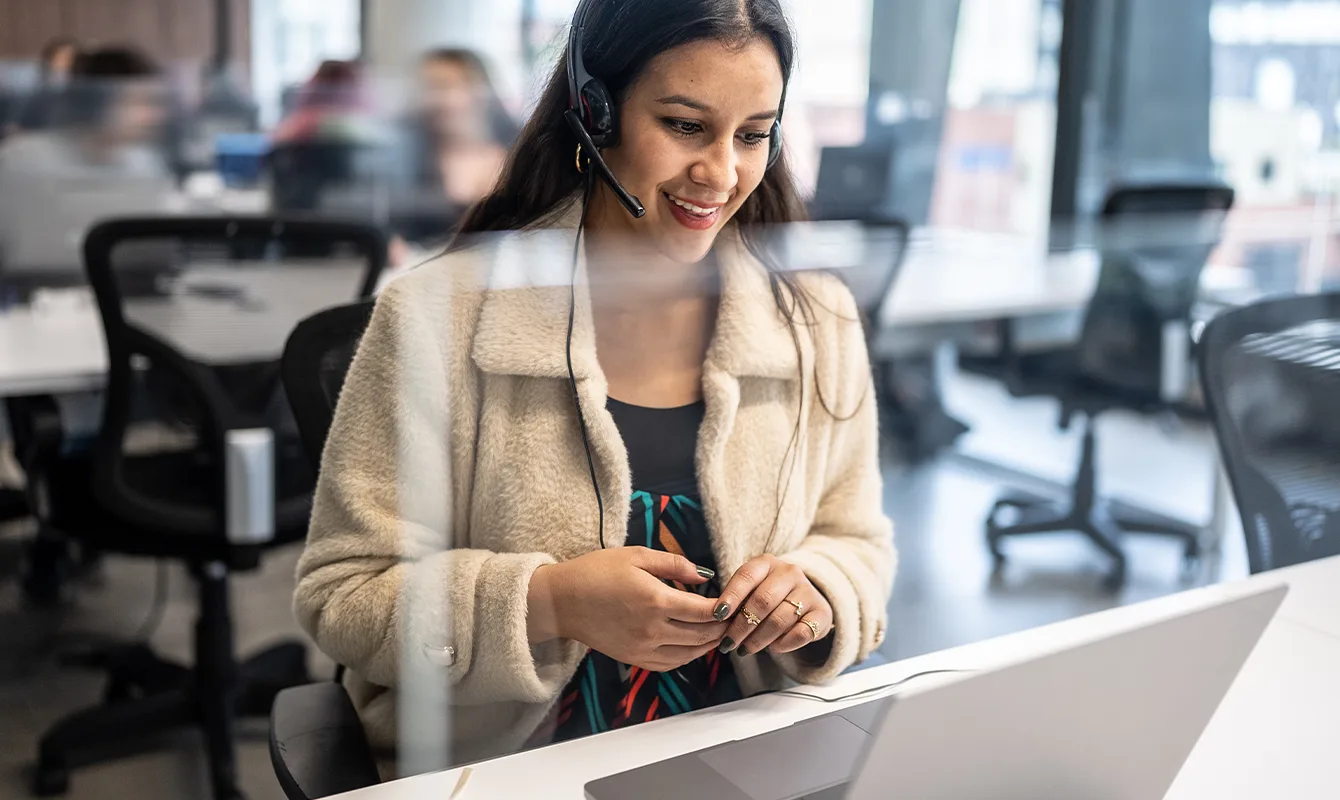 Eine Frau, die ein Headset und eine helle Jacke trägt, sitzt an einem Schreibtisch mit einem Laptop. Sie arbeitet in einem Büro, und im Hintergrund sind weitere Personen verschwommen sichtbar.