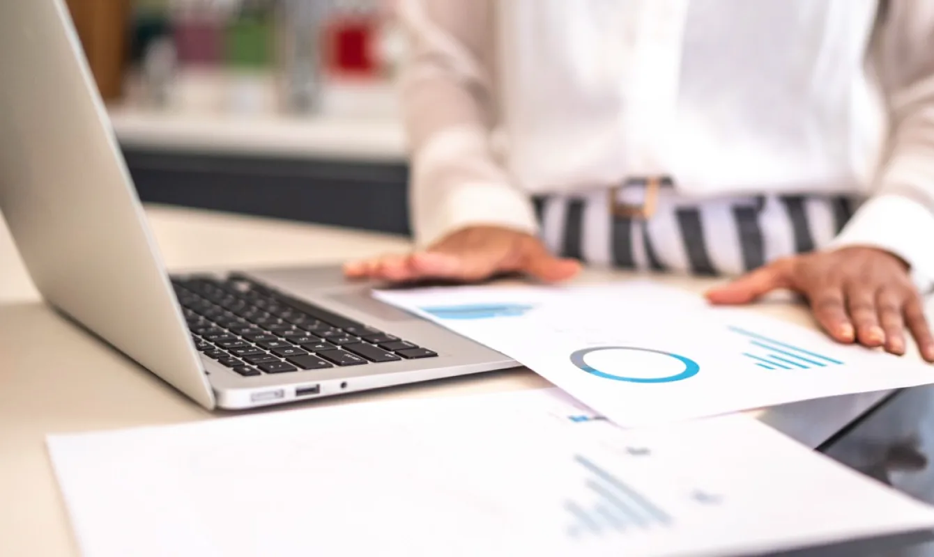 Person standing at a desk, looking at graphs and charts on printed papers, with an open laptop in front of them.