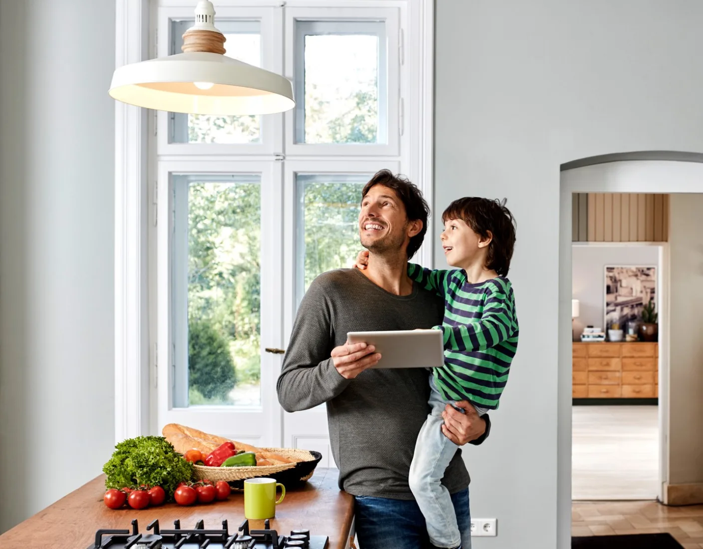 A man holding a tablet in one hand and a child in the other stands by a kitchen counter with fresh vegetables. They are both smiling and looking up towards a light fixture.