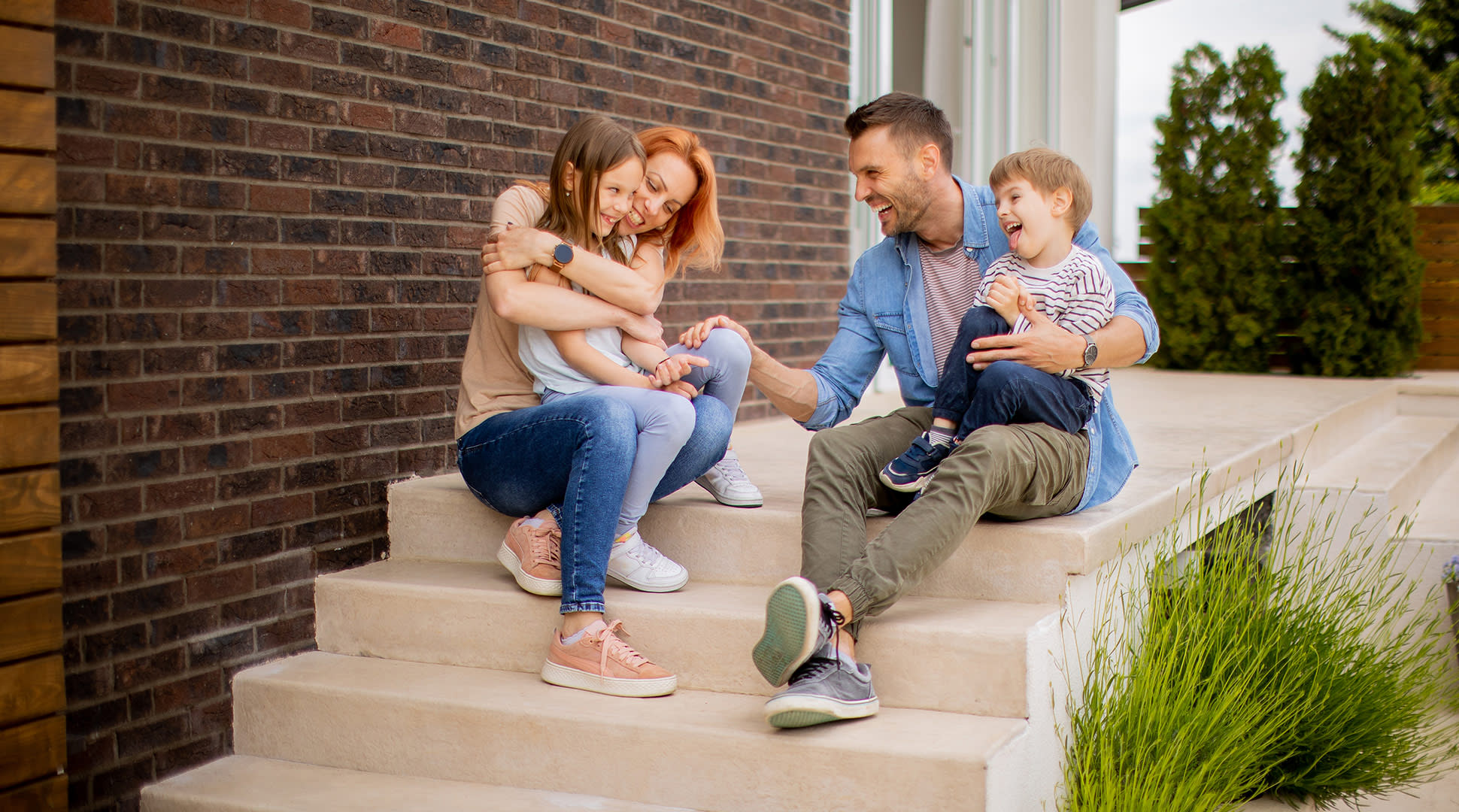 A family of four sits together on the steps of their home, enjoying a sunny day and sharing smiles and laughter.