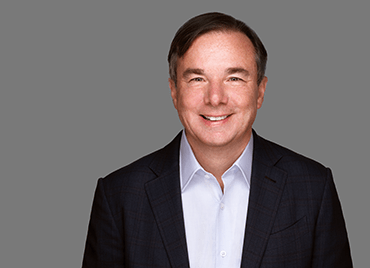 Mike Capone, CEO at Qlik has short brown hair and is wearing a white shirt and dark blazer, smiling against a plain grey background.