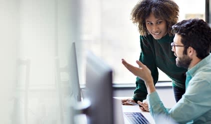 Two colleagues engage in a discussion at a computer workstation, with one pointing at the screen while the other listens.