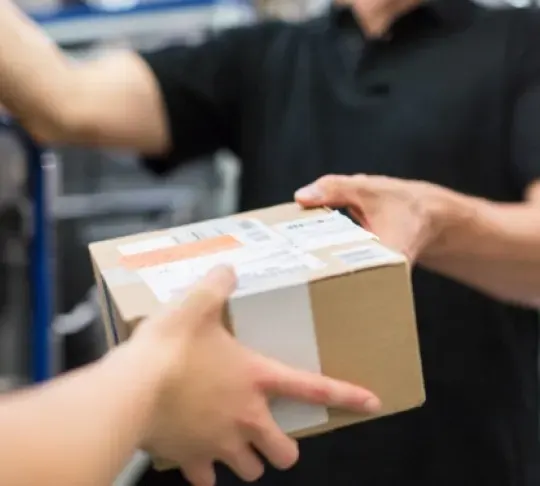 A person in a black shirt hands a cardboard package to another individual in an indoor setting.