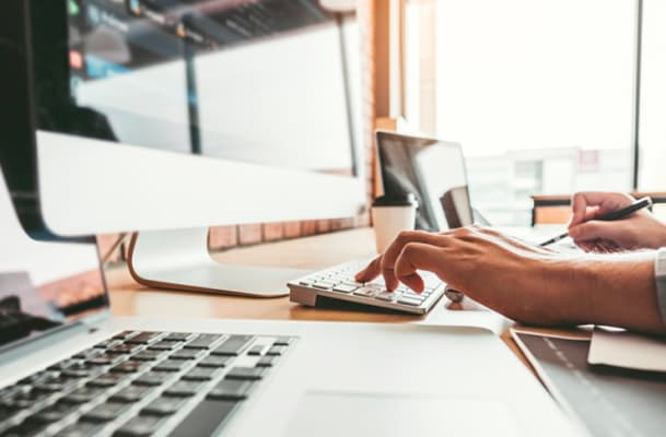 Pessoa digitando em um teclado em frente a várias telas em um escritório bem-iluminado. Um caderno e uma caneta estão próximos a ela.