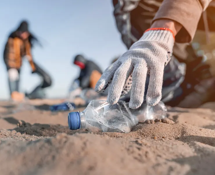 Uma mão com luva pega uma garrafa de plástico na areia da praia, enquanto outras pessoas ao fundo participam da limpeza.