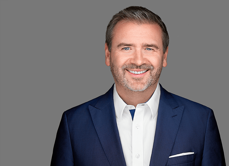 James Fisher, Chief Strategy Officer with short brown hair and beard, wearing a dark blue suit jacket over a white dress shirt, stands smiling in front of a gray background.